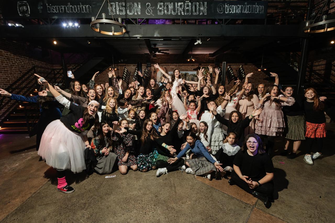 Wide group celebration on the dance floor with many guests jumping and waving their arms, and the couple cheering at the center under warm reception lighting. Bat Mitzvah photography at Bison & Bourbon, Brooklyn, NY.