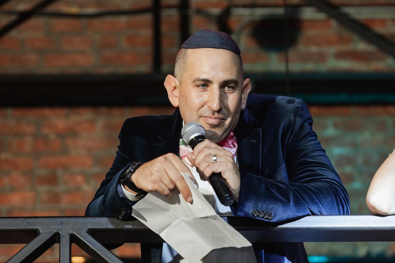 Close-up of a seated emcee holding a microphone and speaking at an event with a brick wall background. Bat Mitzvah photography at Bison & Bourbon, Brooklyn, NY.