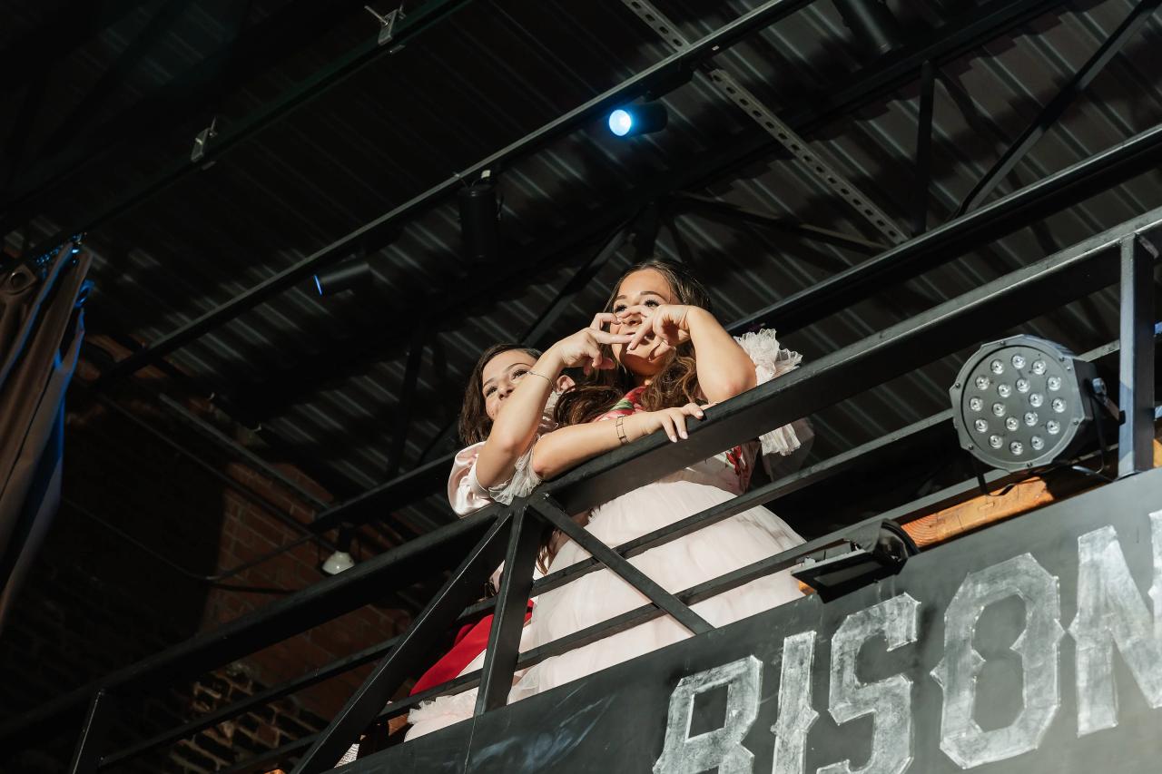 Upward view of a young girl in a light dress posing with her hands making a heart shape during the reception, framed by metal railings and overhead lights. Bat Mitzvah photography at Bison & Bourbon, Brooklyn, NY.