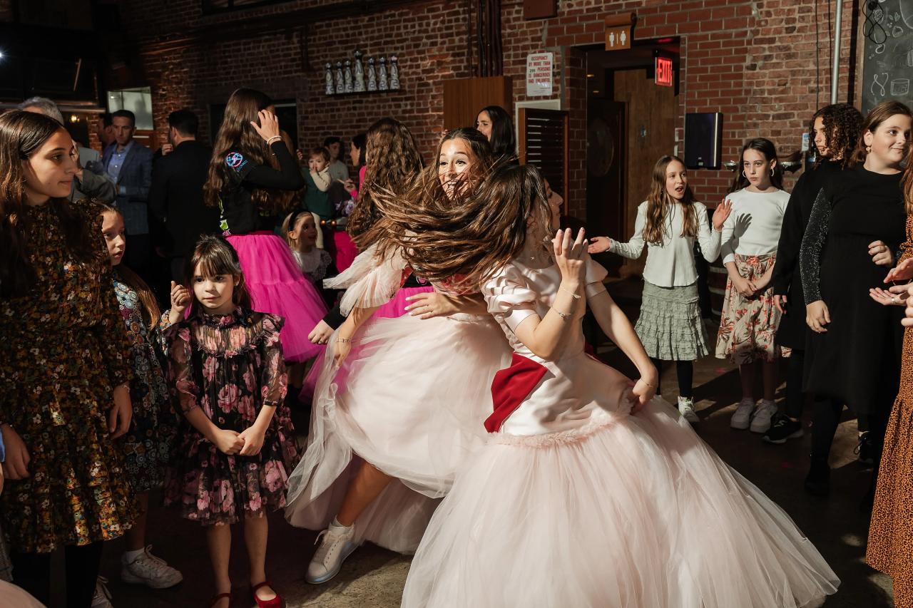 The bride twirls mid-dance on a packed reception floor, hair flying, surrounded by guests and children in warm indoor lighting. Bat Mitzvah photography at Bison & Bourbon, Brooklyn, NY.
