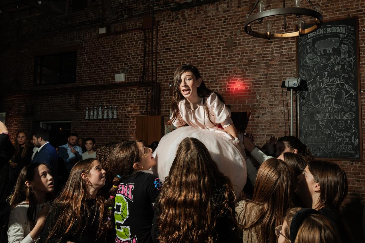 Bride standing above a crowd, gesturing and singing at a reception with exposed brick walls and guests watching closely. Bat Mitzvah photography at Bison & Bourbon, Brooklyn, NY.
