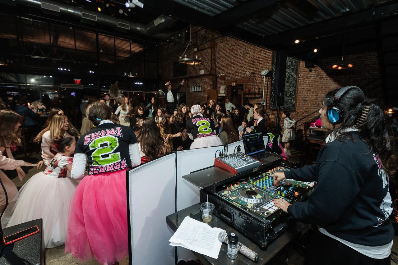 Wedding reception dance floor with a DJ performing at a booth and guests moving nearby, including children in bright festive outfits. Bat Mitzvah photography at Bison & Bourbon, Brooklyn, NY.