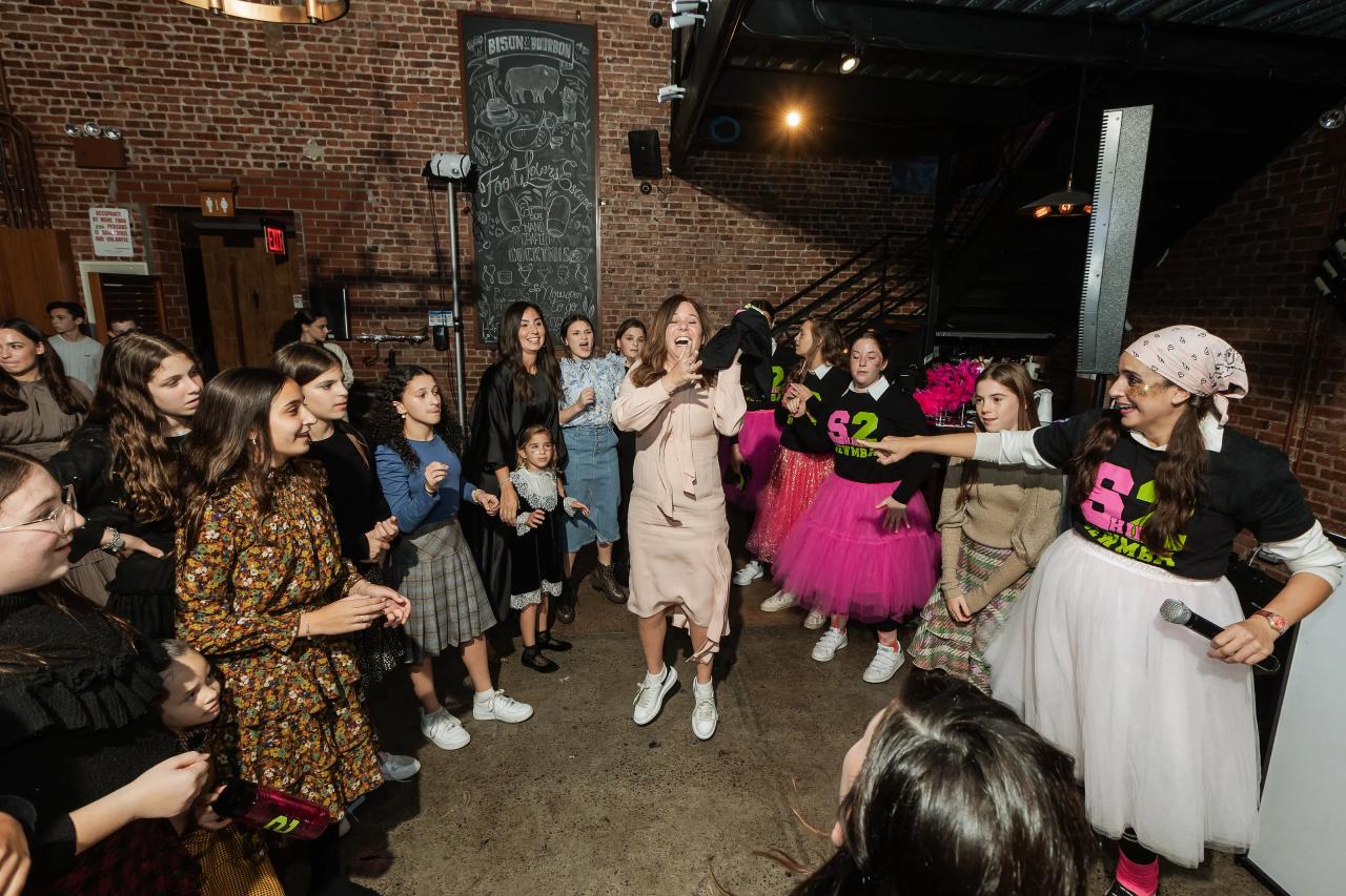 Bride laughing in the center of a lively reception dance floor, surrounded by children and guests, with exposed brick walls and bright stage lighting behind her. Bat Mitzvah photography at Bison & Bourbon, Brooklyn, NY.