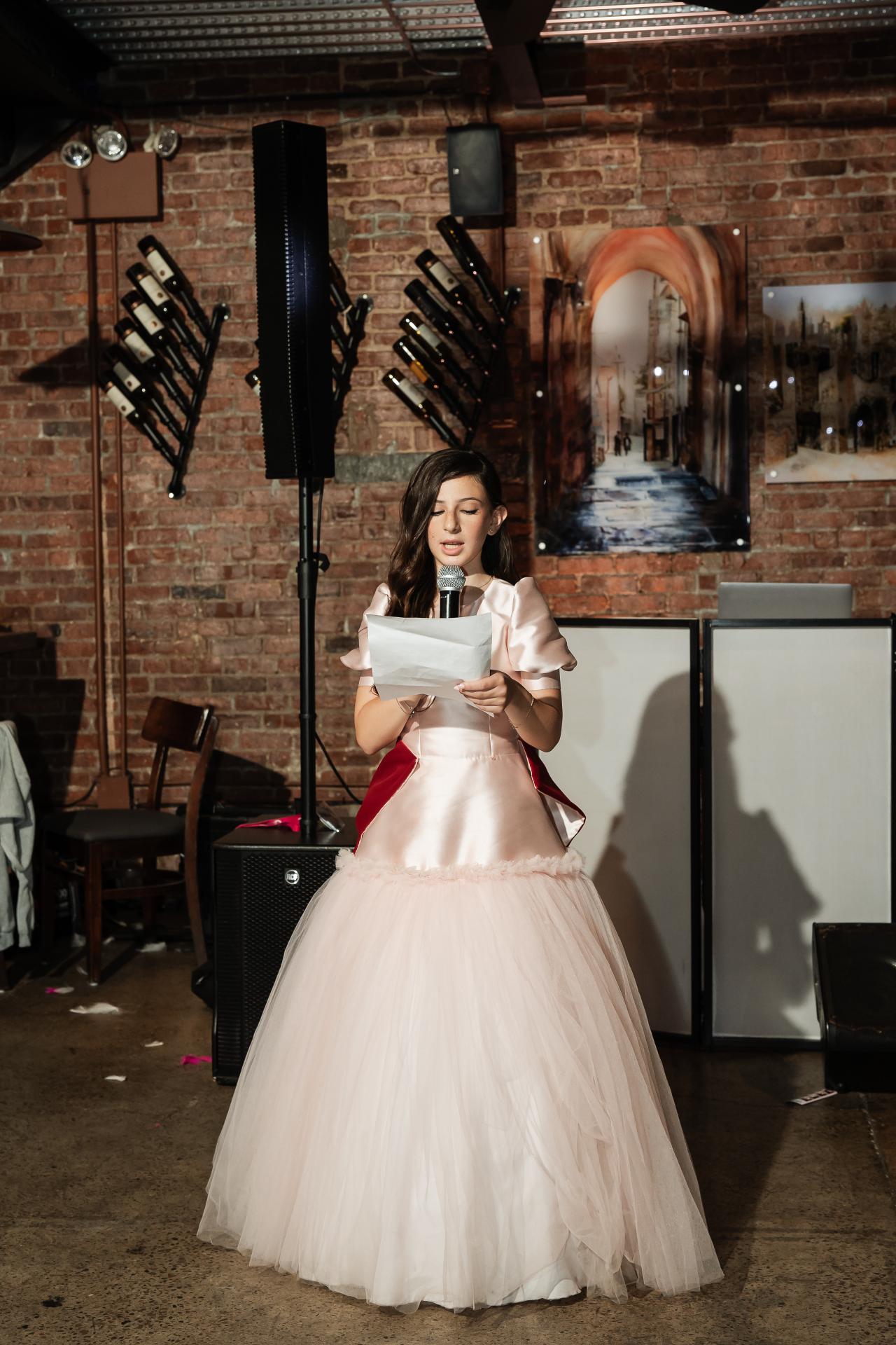 Bat mitzvah girl on stage holding notes and speaking into a microphone in a brick-walled venue. Bat Mitzvah photography at Bison & Bourbon, Brooklyn, NY.