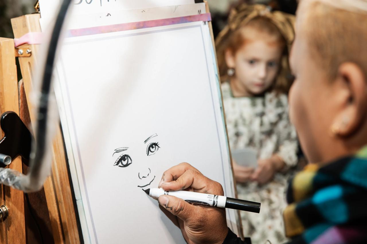 Close-up of a hand using a marker to draw facial details on a white board at a wedding event, with softly blurred guests in the background. Bat Mitzvah photography at Bison & Bourbon, Brooklyn, NY.