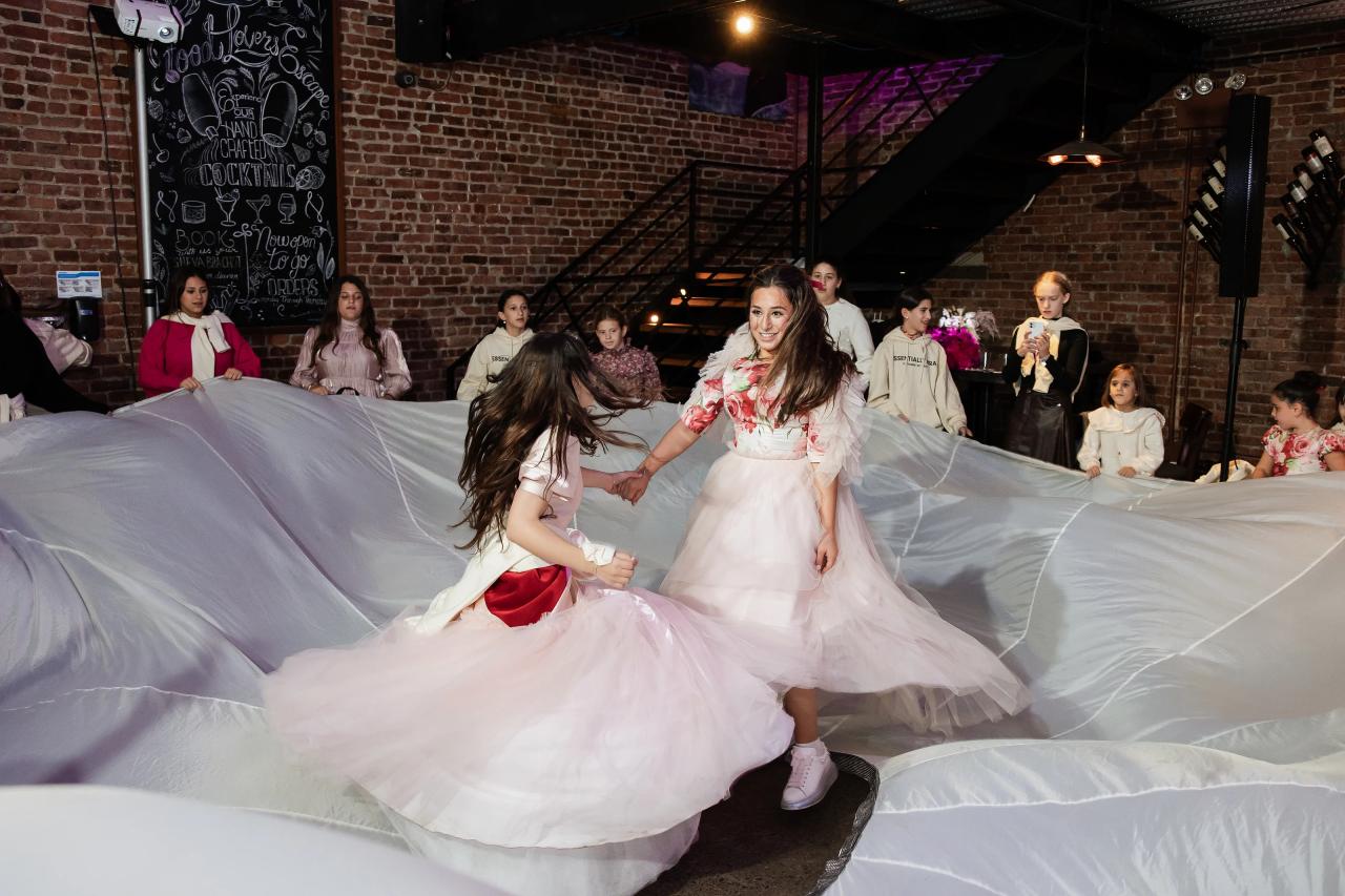 Bride smiling at center while children move beneath and around a large white sheer fabric drape on the dance floor in an indoor brick hall. Bat Mitzvah photography at Bison & Bourbon, Brooklyn, NY.