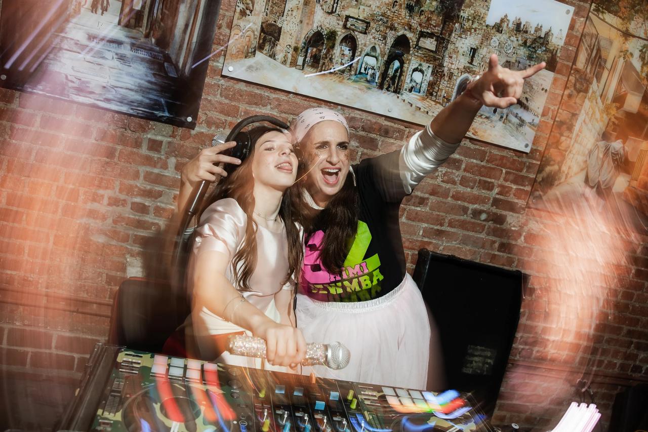 Joyful reception moment with a bride and groom laughing and cheering at a DJ booth, hands raised, brick wall background, and colorful light streaks. Bat Mitzvah photography at Bison & Bourbon, Brooklyn, NY.