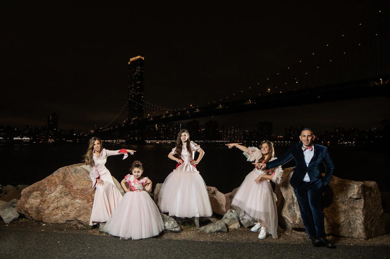 Night waterfront portrait of a couple and children in formal attire, posing with arms outstretched in front of a lit city skyline. Bat Mitzvah photography at Bison & Bourbon, Brooklyn, NY.