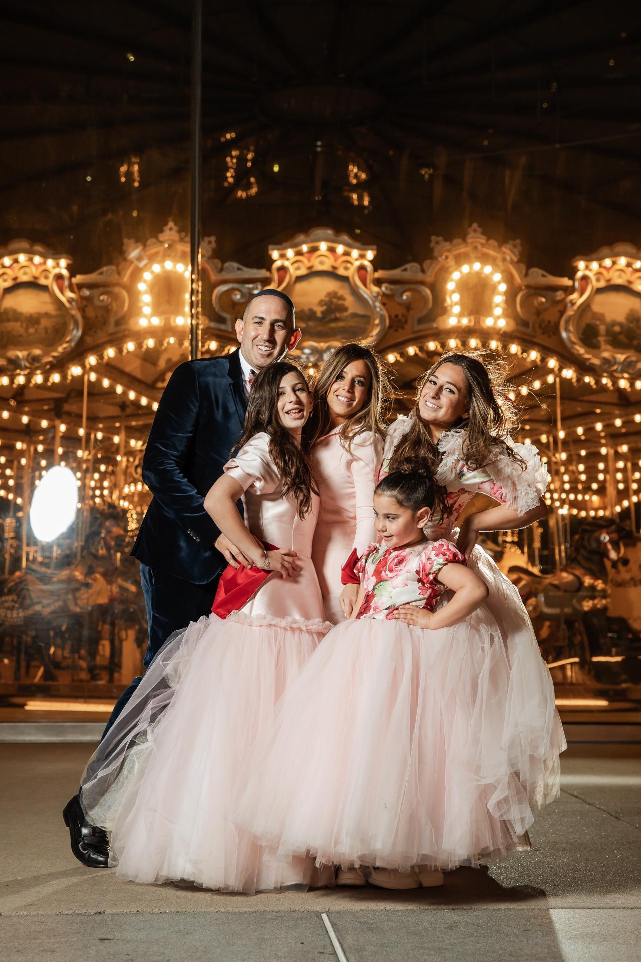 Smiling family group portrait on a wedding day in a warmly lit ballroom with ornate golden chairs and chandelier lights behind them. Bat Mitzvah photography at Bison & Bourbon, Brooklyn, NY.