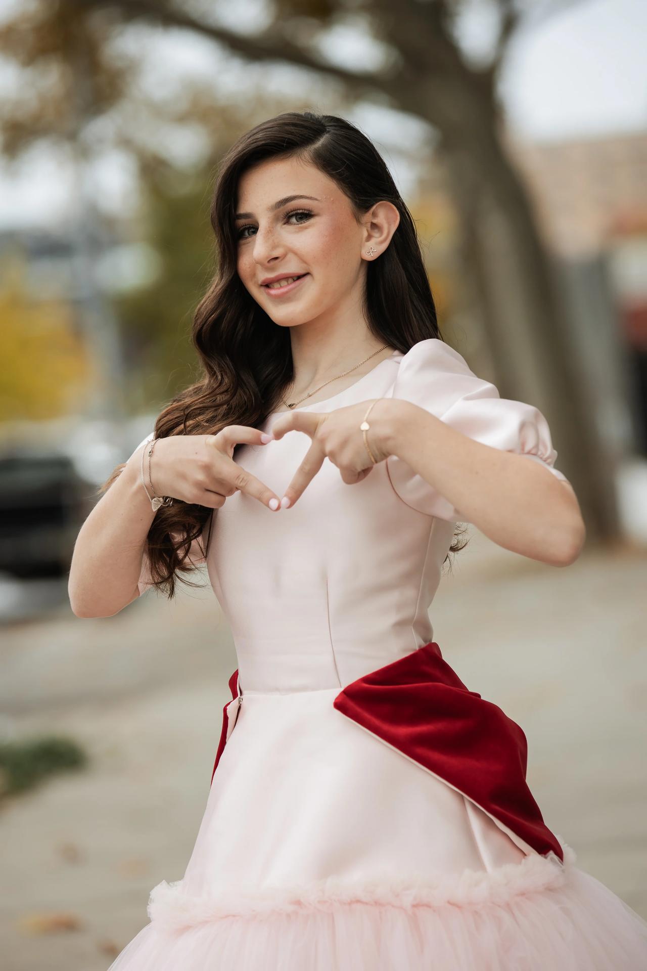 Mitzvah portrait of a girl in a light dress and red sash making a heart gesture with her hands outdoors, softly blurred background. Bat Mitzvah photography at Bison & Bourbon, Brooklyn, NY.