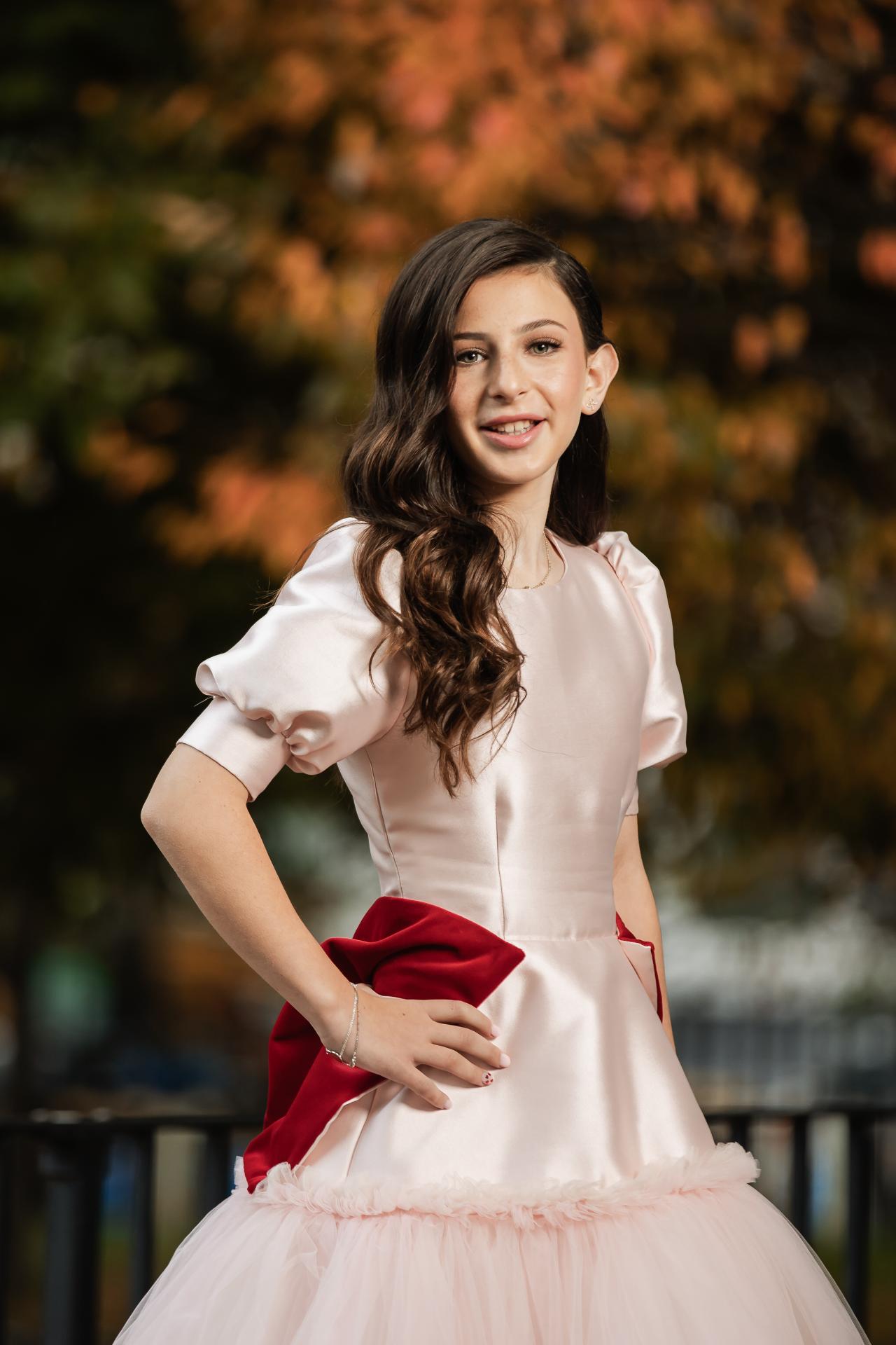Close-up outdoor portrait of a girl in a light satin bat mitzvah dress with a deep red sash, smiling softly against a warm blurred background of autumn trees. Bat Mitzvah photography at Bison & Bourbon, Brooklyn, NY.