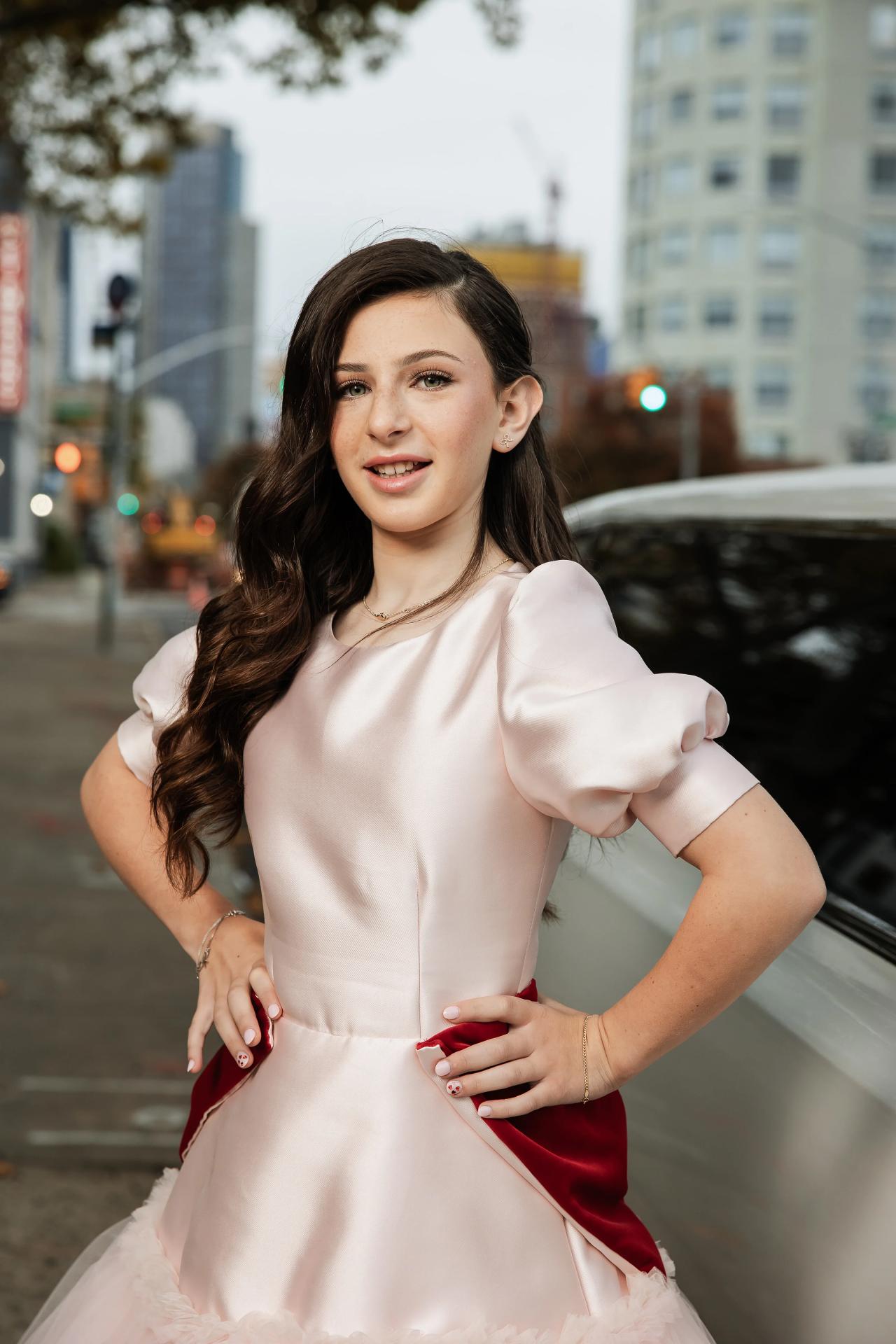 Elegant celebrant portrait outdoors in front of city buildings, wearing a light dress with a deep red satin sash and a confident expression. Bat Mitzvah photography at Bison & Bourbon, Brooklyn, NY.