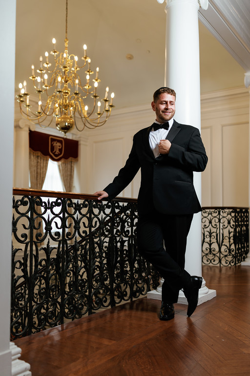 Groom in a black tuxedo and bow tie smiling while standing on a stair landing near an ornate railing and chandelier.