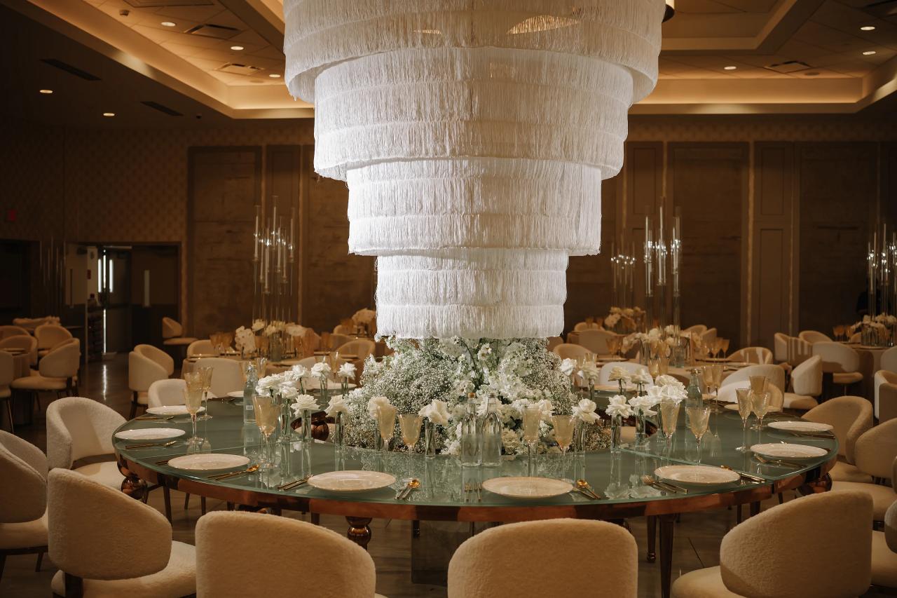 Wide view of a reception ballroom with a large hanging chandelier centerpiece and styled round tables with white linens, gold glassware, and floral arrangements. Bar Mitzvah photography at Hilton Garden Inn Springfield, Springfield, NJ.