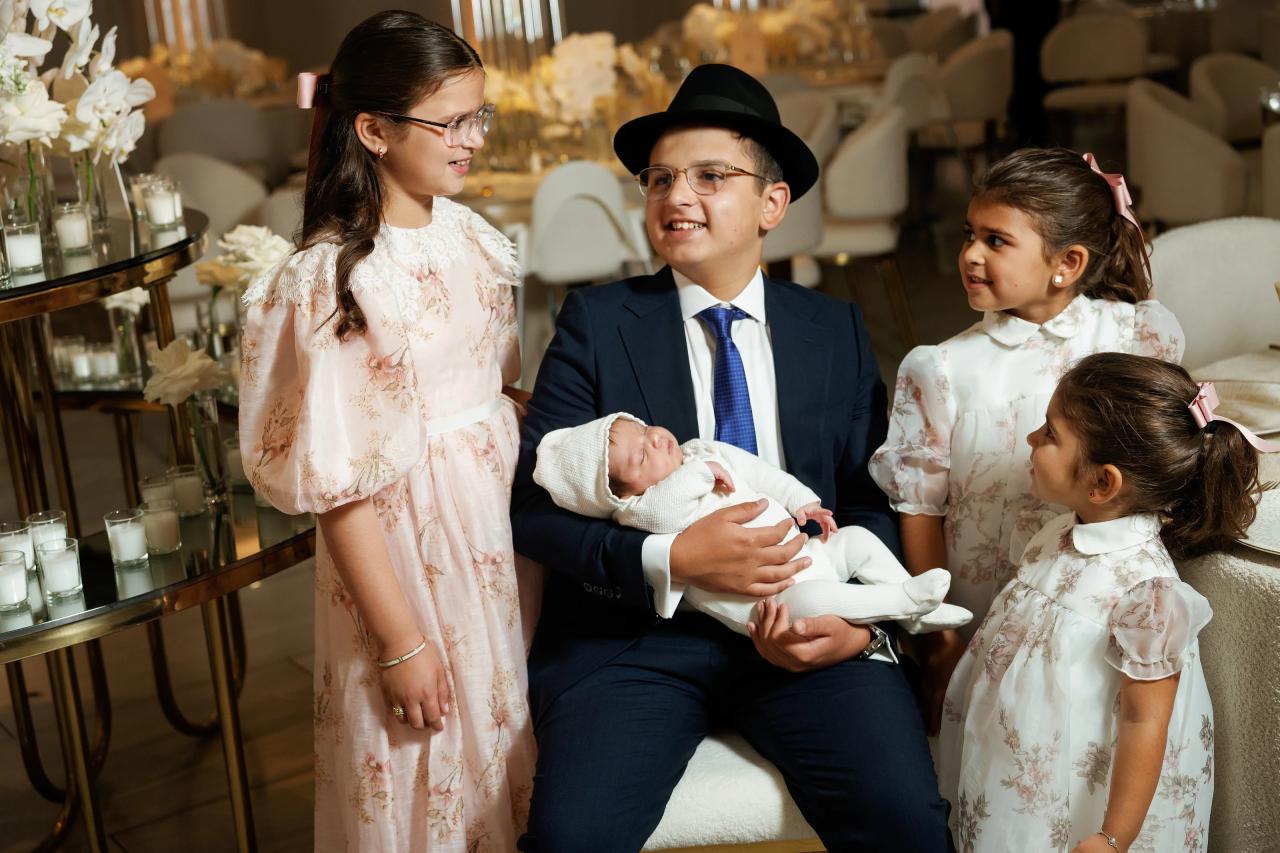 Parents seated with a newborn while four children in coordinated outfits smile during a bar mitzvah family moment. Bar Mitzvah photography at Hilton Garden Inn Springfield, Springfield, NJ.