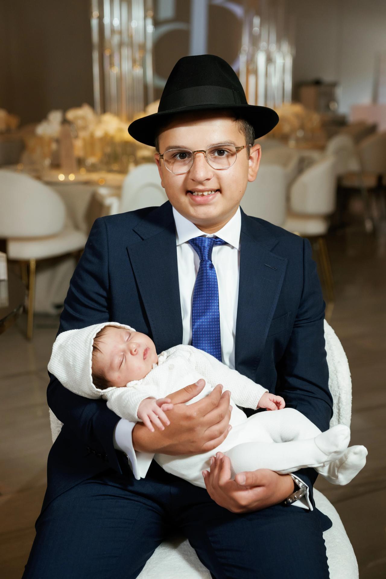 A young man in a black hat and suit holding a swaddled newborn for a warm indoor simcha portrait, with gold event décor softly blurred in the background. Bar Mitzvah photography at Hilton Garden Inn Springfield, Springfield, NJ.
