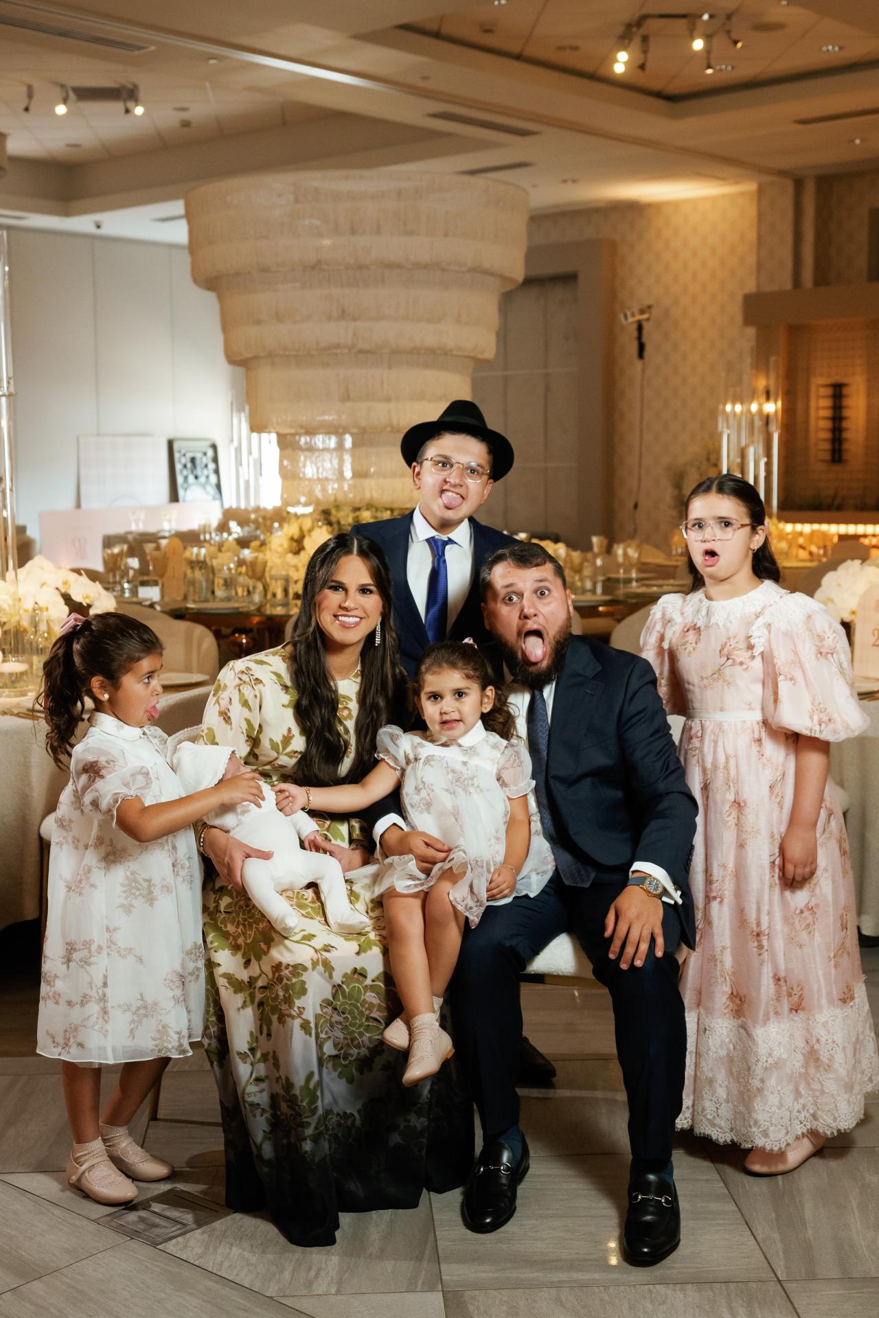 Warm, indoor family group photo at a Jewish celebration, with parents in formal traditional attire and children in coordinated dresses, captured with playful expressions. Bar Mitzvah photography at Hilton Garden Inn Springfield, Springfield, NJ.