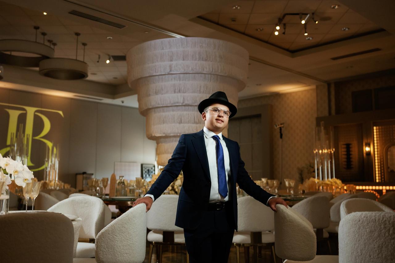 Young guest in formalwear wearing a hat and blue tie standing between lounge chairs in a grand banquet hall, arms resting on the chair backs, with a large round centerpiece in the background. Bar Mitzvah photography at Hilton Garden Inn Springfield, Springfield, NJ.