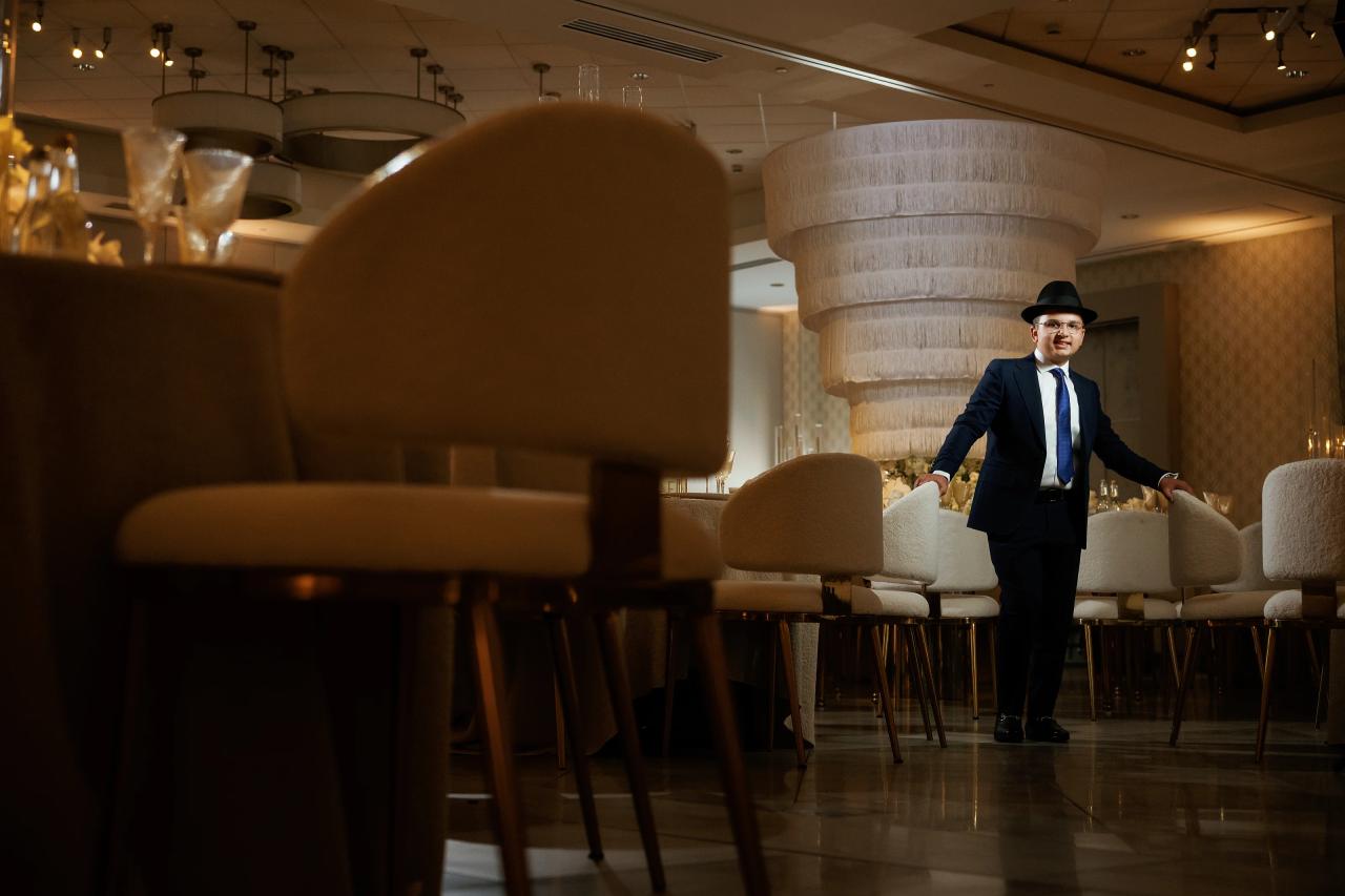 A formally dressed young man in a black hat and suit standing in a warmly lit event hall near chairs, with soft focus decor behind him. Bar Mitzvah photography at Hilton Garden Inn Springfield, Springfield, NJ.