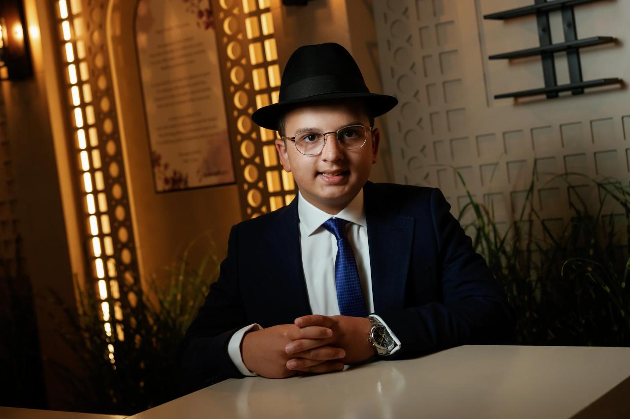 Framed portrait of a young man in a dark hat, glasses, and formal suit seated at a table with warm side lighting in the background. Bar Mitzvah photography at Hilton Garden Inn Springfield, Springfield, NJ.