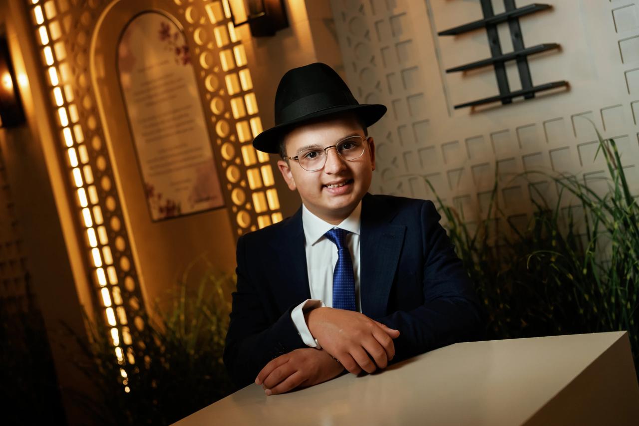 Smiling young man in a fedora and suit seated at a table, framed by warm architectural lighting and decorative wall panels. Bar Mitzvah photography at Hilton Garden Inn Springfield, Springfield, NJ.