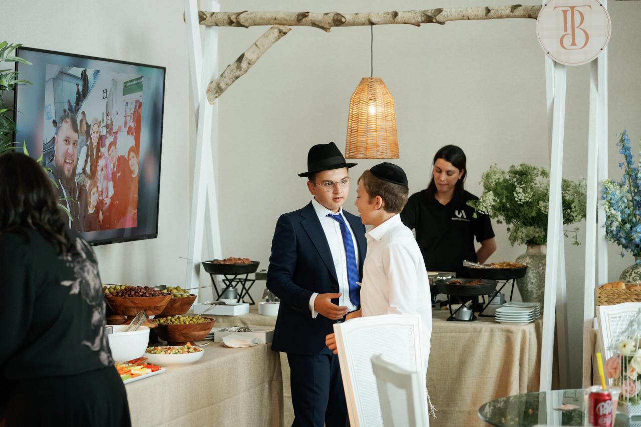 Two young men in formal attire standing near a decorated buffet table, facing each other and speaking during a simcha. Bar Mitzvah photography at Hilton Garden Inn Springfield, Springfield, NJ.