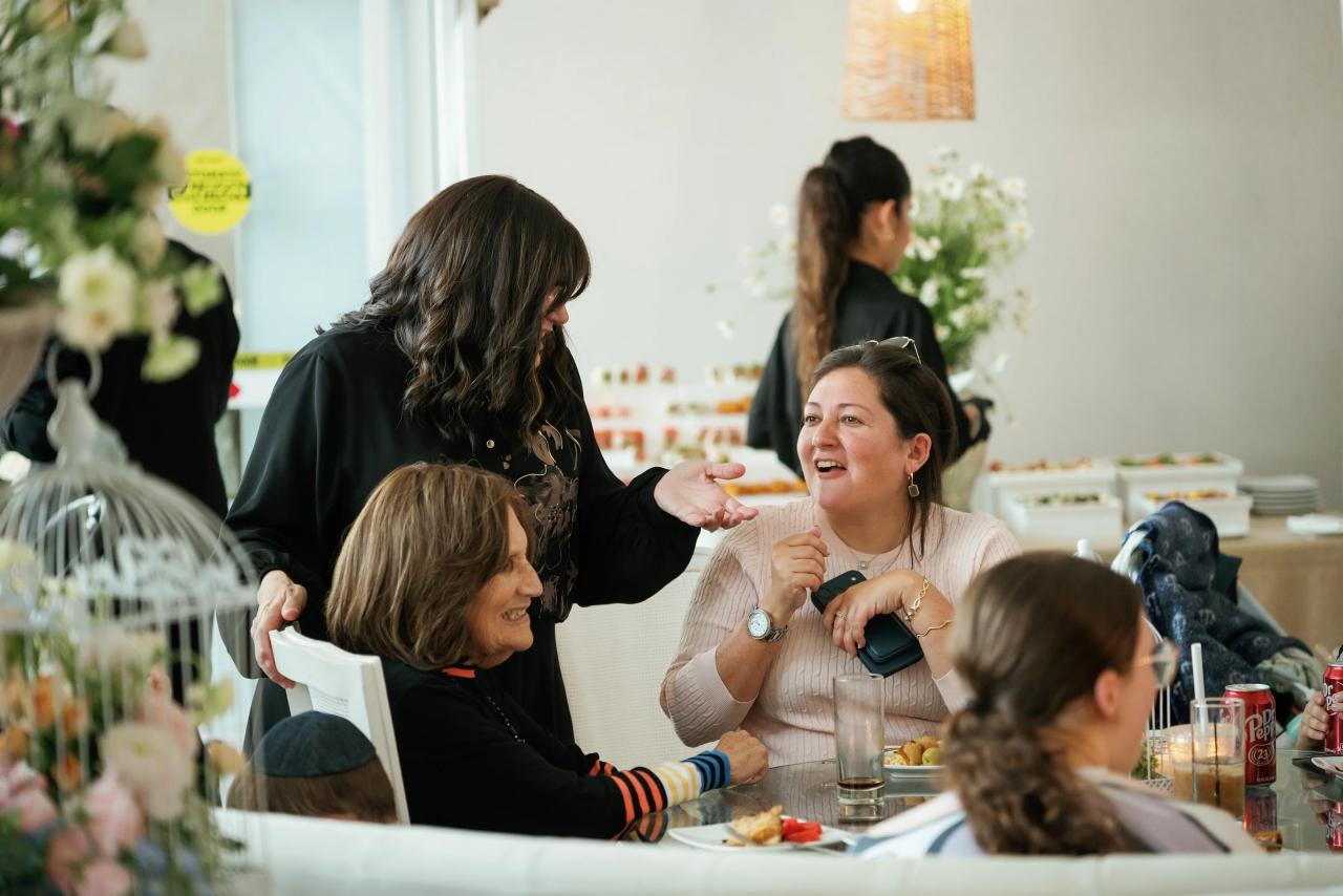 Guests laugh and chat at a bar mitzvah table, with drinks, food and a floral centerpiece in the foreground. Bar Mitzvah photography at Hilton Garden Inn Springfield, Springfield, NJ.