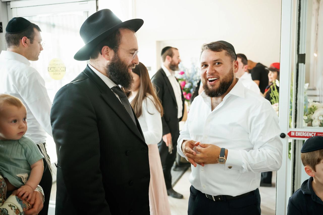 A smiling guest in a black hat and dark suit laughs while greeting another guest in a bright indoor corridor. Bar Mitzvah photography at Hilton Garden Inn Springfield, Springfield, NJ.