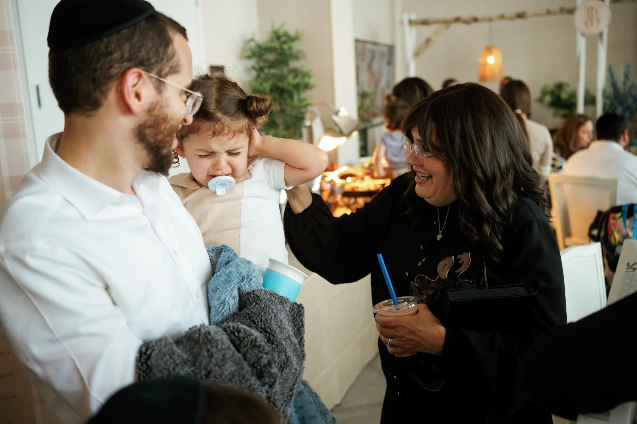 Smiling parents cradle a baby at a reception table while guests sit in the background; warm candlelight and casual celebration atmosphere. Bar Mitzvah photography at Hilton Garden Inn Springfield, Springfield, NJ.