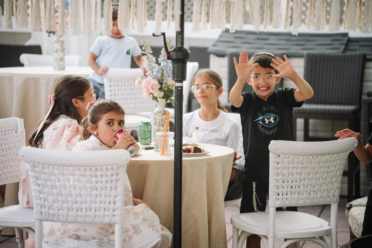 Children smiling and gesturing joyfully at a reception table with a floral centerpiece and tassel décor in the background. Bar Mitzvah photography at Hilton Garden Inn Springfield, Springfield, NJ.