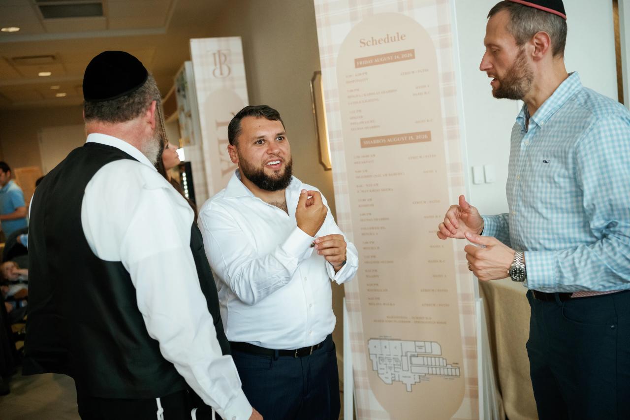 Candid event moment: a smiling man gestures near a standing schedule board as others react and converse in a lobby. Bar Mitzvah photography at Hilton Garden Inn Springfield, Springfield, NJ.