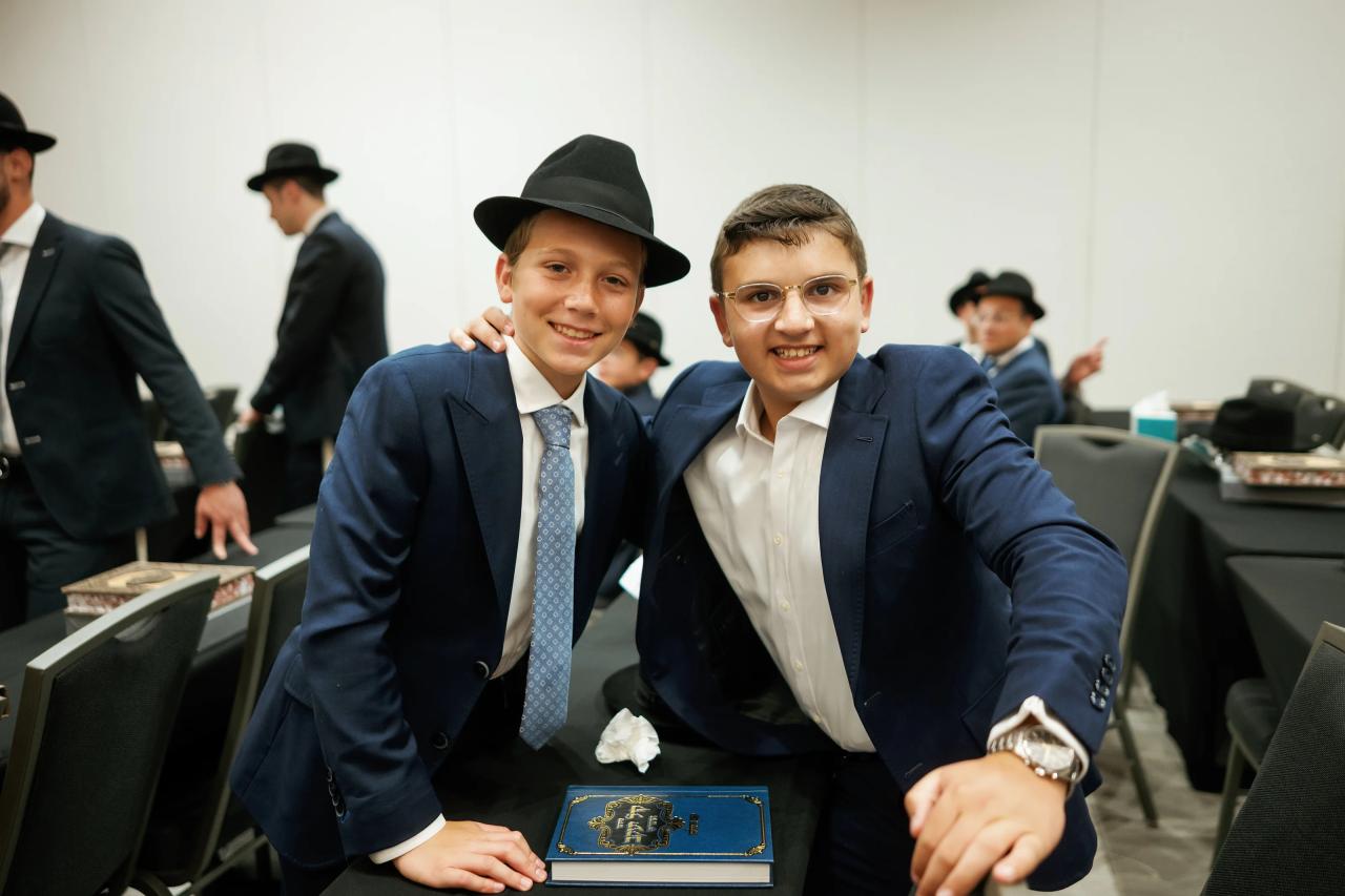 Two young men in traditional attire smiling closely at the table during an event, with other guests seated in the background. Bar Mitzvah photography at Hilton Garden Inn Springfield, Springfield, NJ.