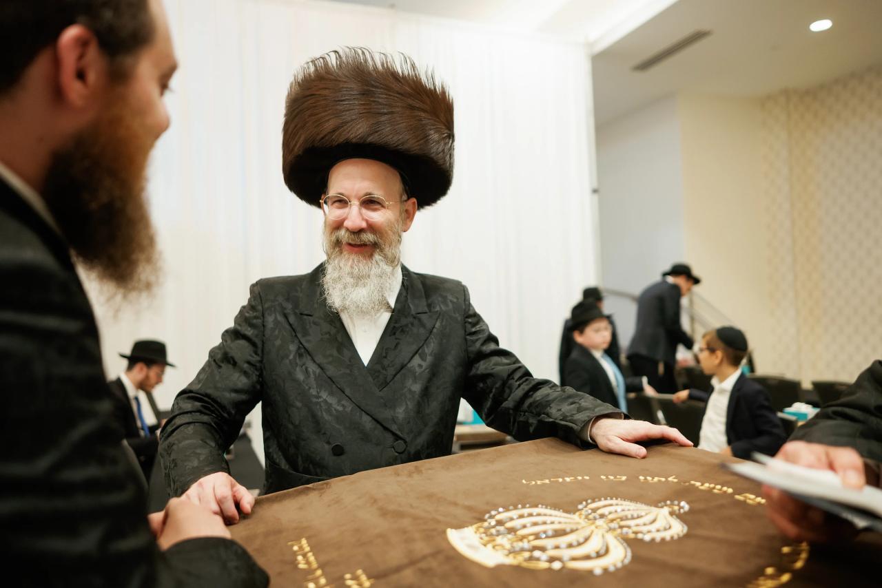 An older man in a black hat sits behind a decorated reception table during the bar mitzvah. Bar Mitzvah photography at Hilton Garden Inn Springfield, Springfield, NJ.