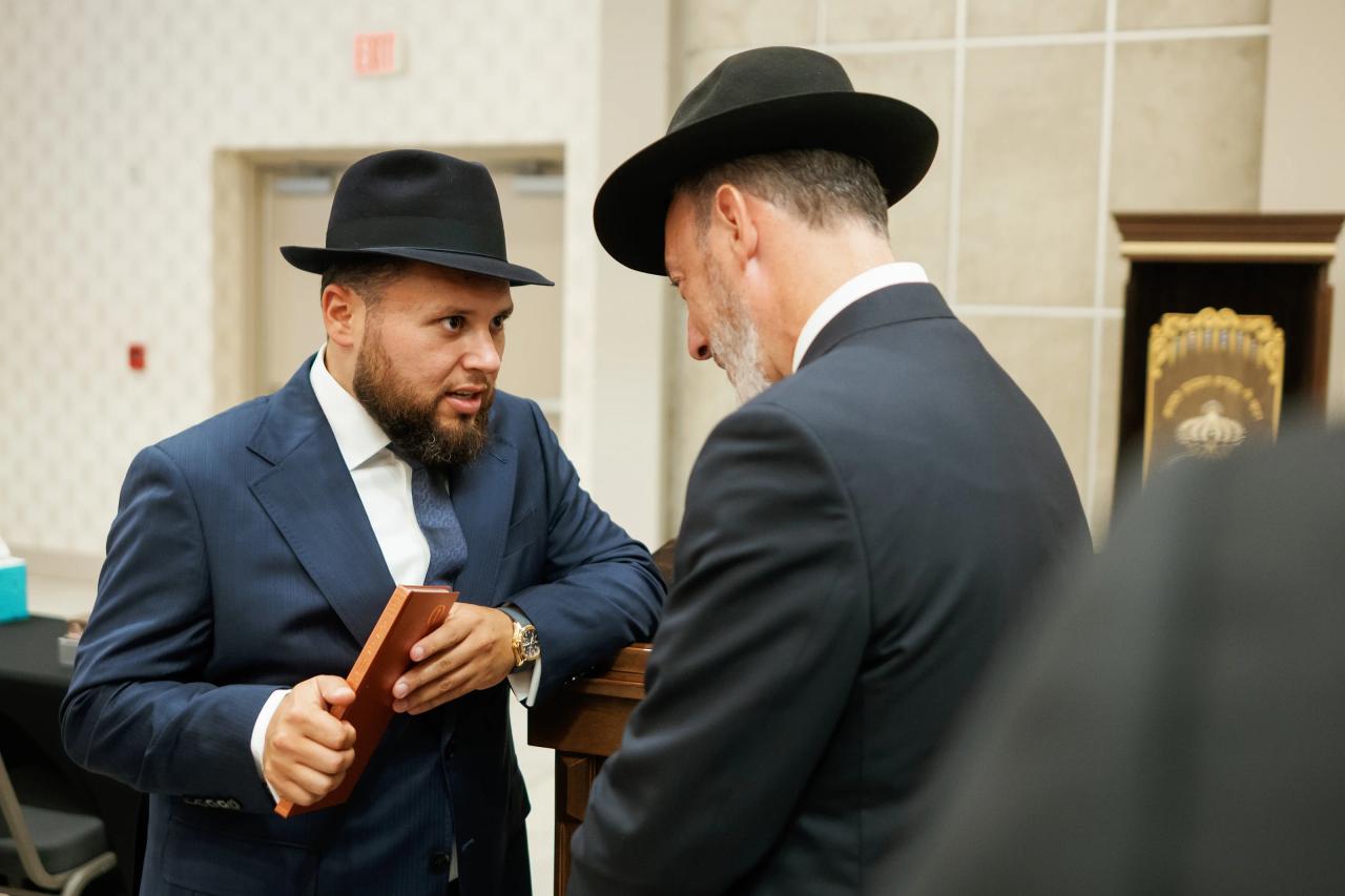 Two men in formal suits facing each other closely during an indoor ceremony, one holding a small book-like item while speaking. Bar Mitzvah photography at Hilton Garden Inn Springfield, Springfield, NJ.