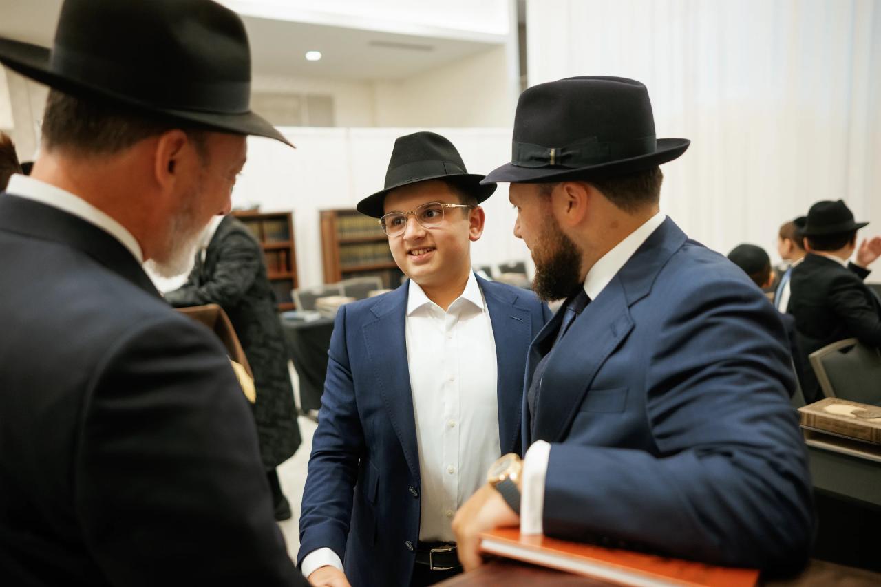 A young guest in a suit and hat shares a smile with an older man seated at the bar mitzvah reception. Bar Mitzvah photography at Hilton Garden Inn Springfield, Springfield, NJ.