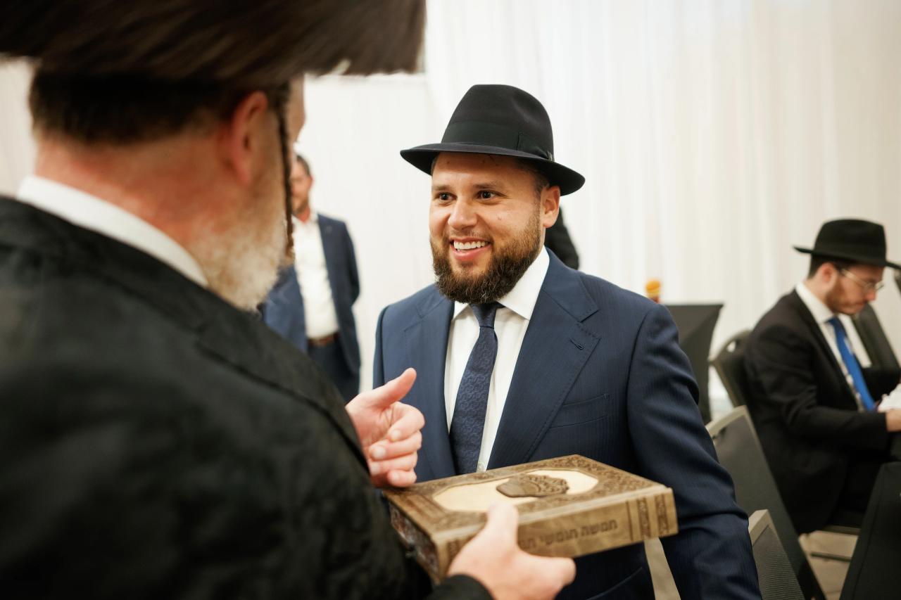 A smiling guest in a black hat and suit, with a presentation held in the foreground and a companion in soft focus. Bar Mitzvah photography at Hilton Garden Inn Springfield, Springfield, NJ.
