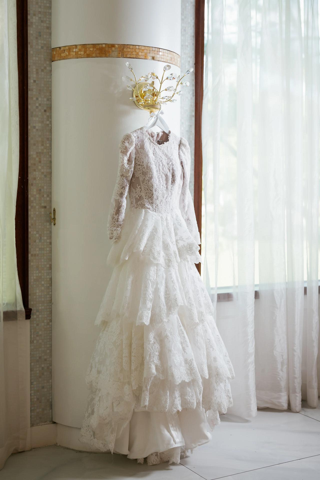 A lace wedding dress hanging beside a bright window and sheer curtains, photographed before the bride gets dressed. Wedding photography at Riverview Ballroom, Cliffside Park, NJ.