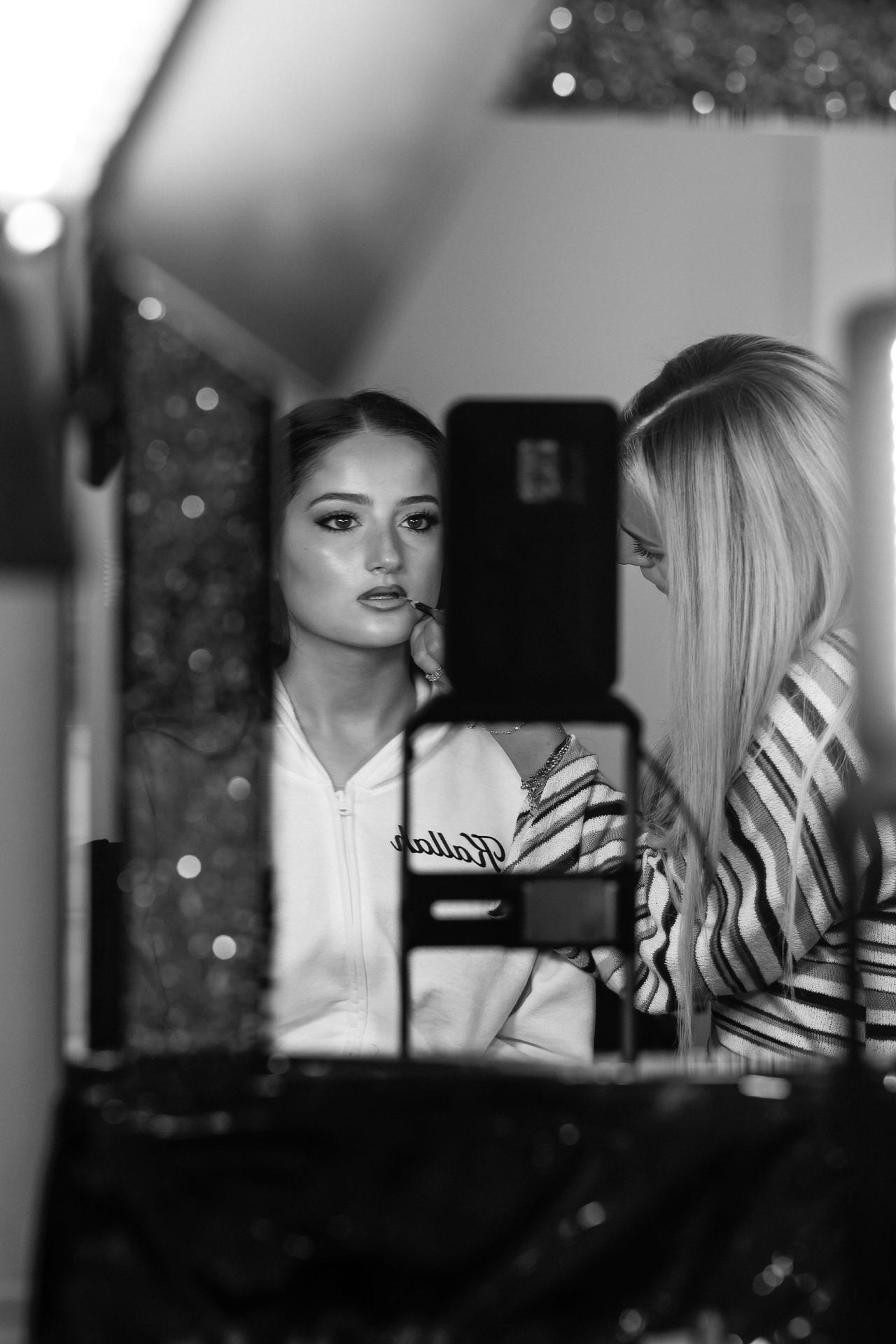 Black and white candid of two women indoors; one looks toward the camera while holding up a smartphone, with soft bokeh lights in the background. Wedding photography at Riverview Ballroom, Cliffside Park, NJ.