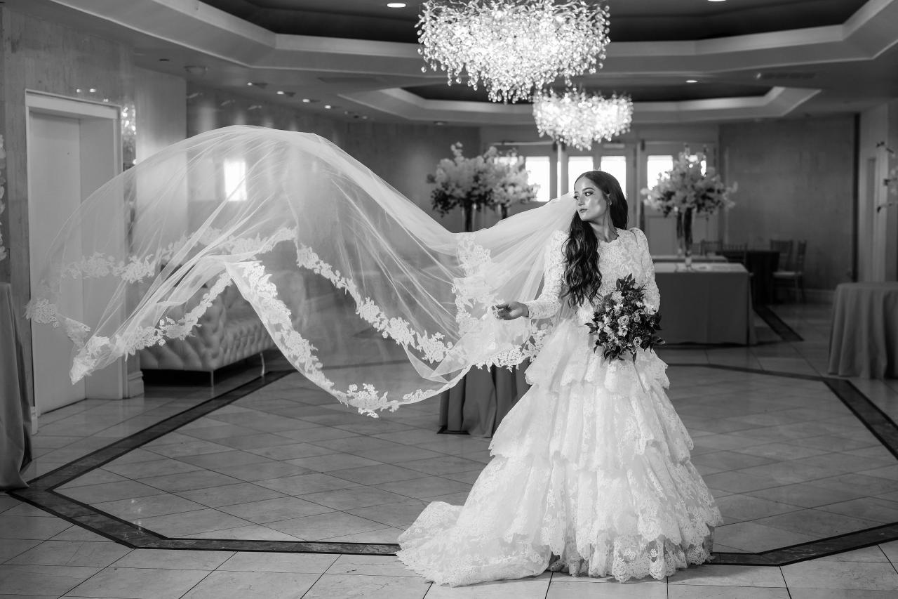 Black-and-white wedding photo of the bride in a lace gown walking with her veil flowing, bouquet in hand, chandelier lights overhead in a reception hall. Wedding photography at Riverview Ballroom, Cliffside Park, NJ.