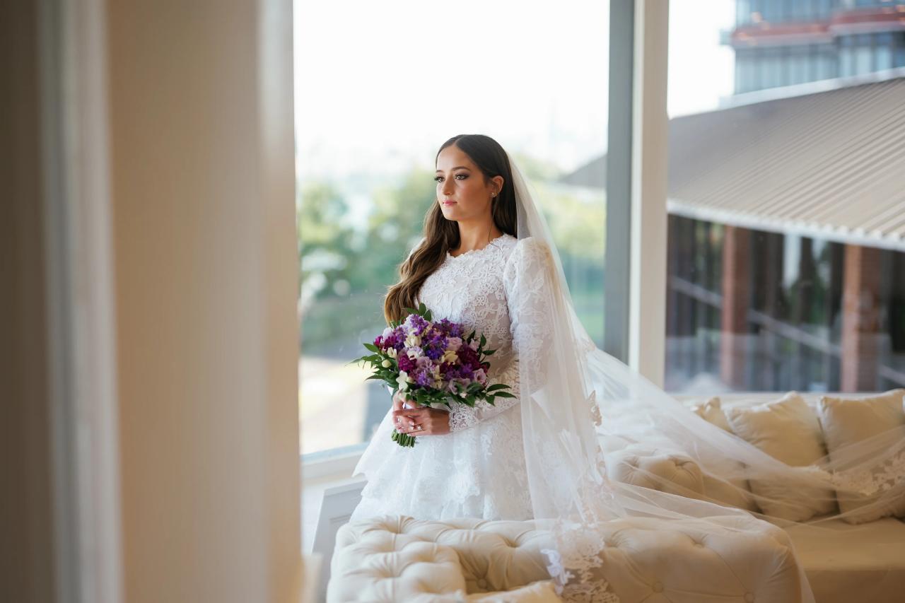 Bride in a lace white wedding dress and veil holding a bouquet of purple and white flowers indoors, framed by white columns and soft daylight. Wedding photography at Riverview Ballroom, Cliffside Park, NJ.