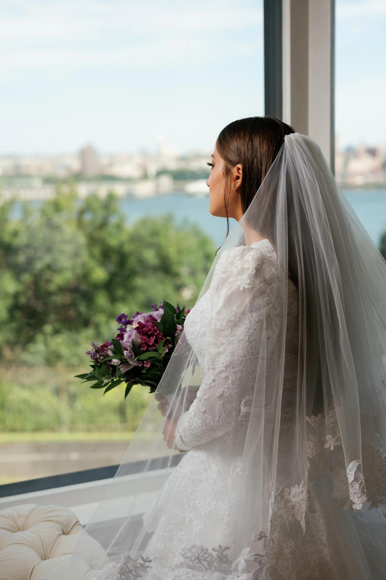 Bride in a white gown and flowing veil in side profile holding a bouquet, with a scenic outdoor view in the background. Wedding photography at Riverview Ballroom, Cliffside Park, NJ.