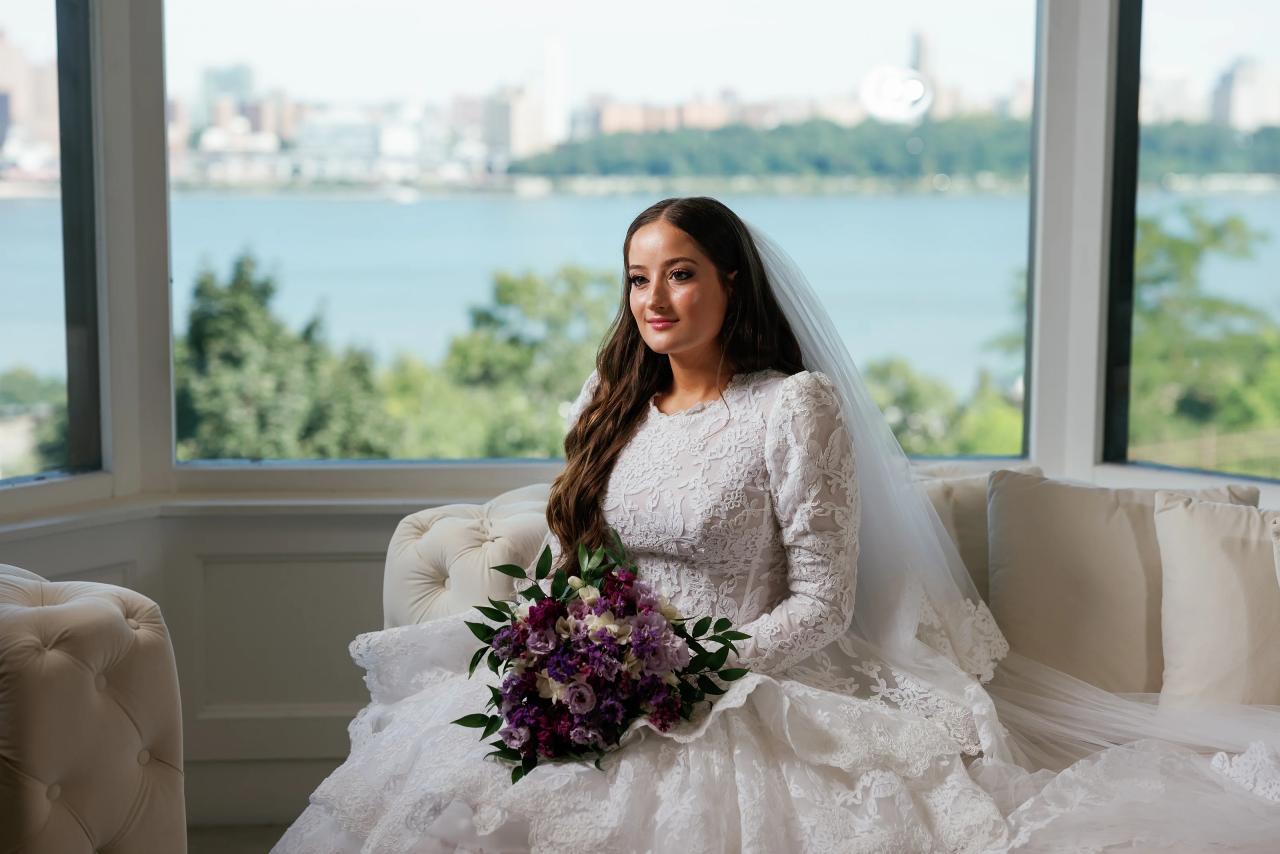 Bride in lace gown and veil seated by a large window holding a bouquet, with a scenic water view in the background. Wedding photography at Riverview Ballroom, Cliffside Park, NJ.