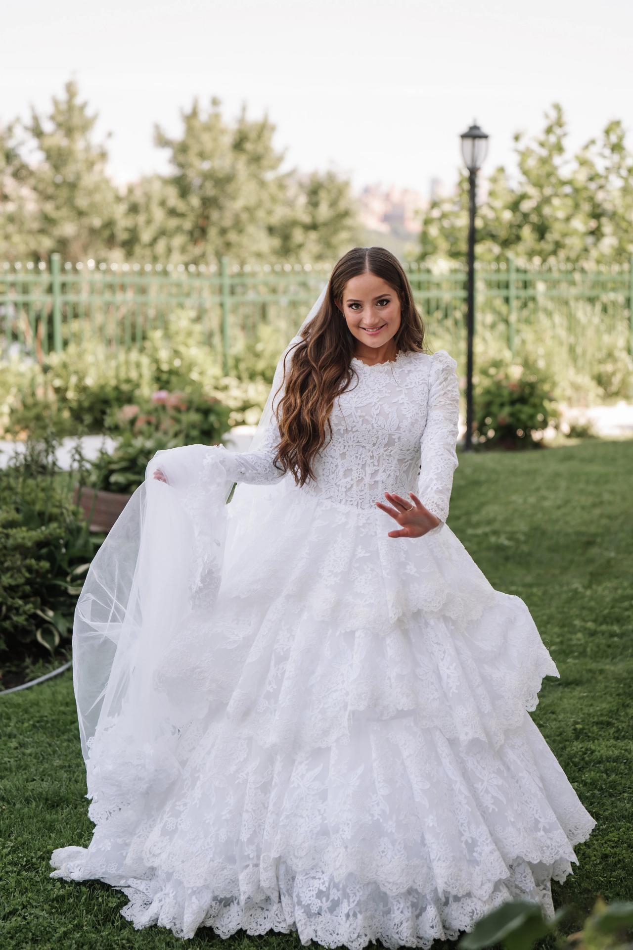 Bride in a white lace wedding gown smiling outdoors in a garden, lifting her skirt/tulle in the sunlight. Wedding photography at Riverview Ballroom, Cliffside Park, NJ.