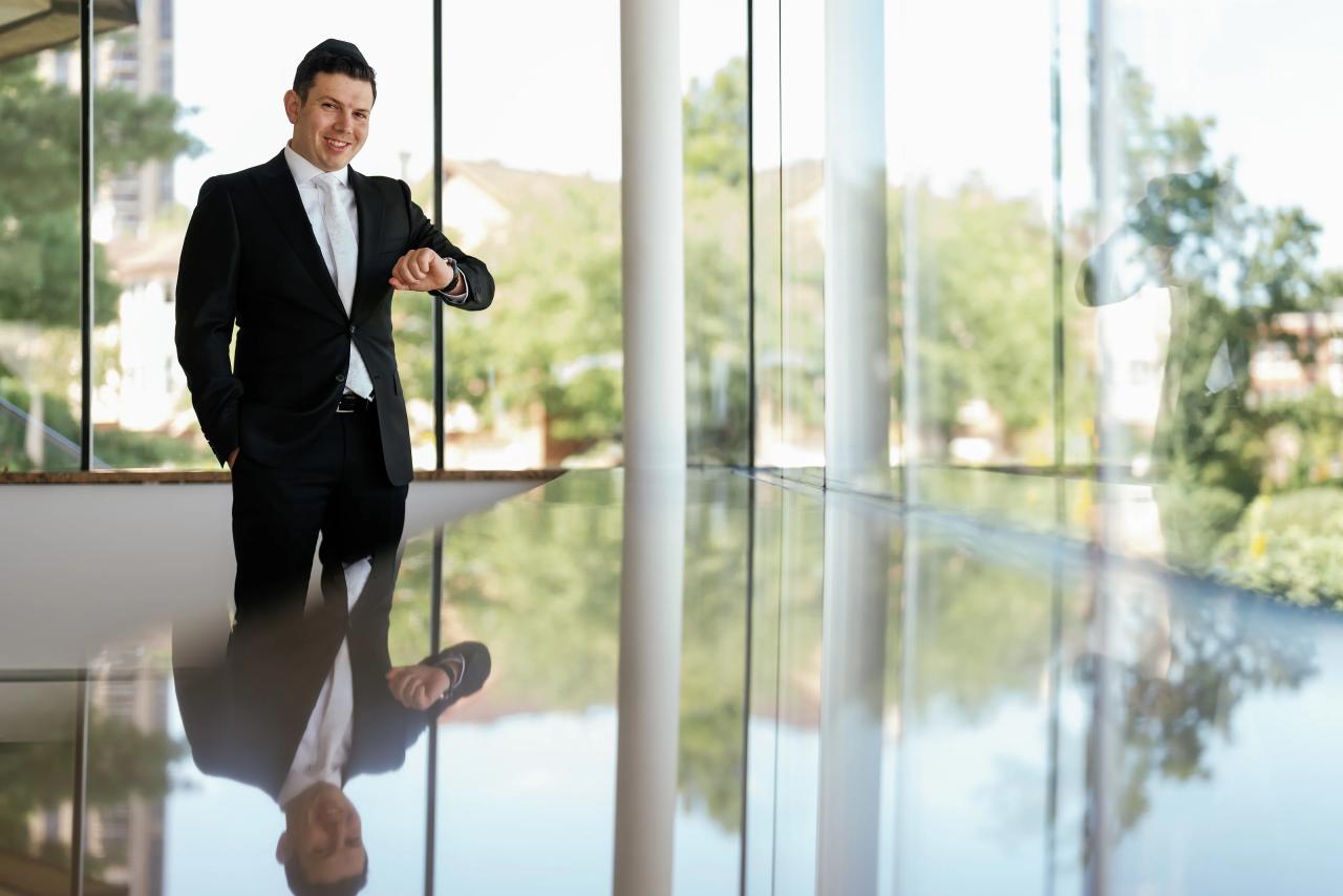 Smiling groom standing near large glass panels with reflections, daytime light in a modern indoor space. Wedding photography at Riverview Ballroom, Cliffside Park, NJ.