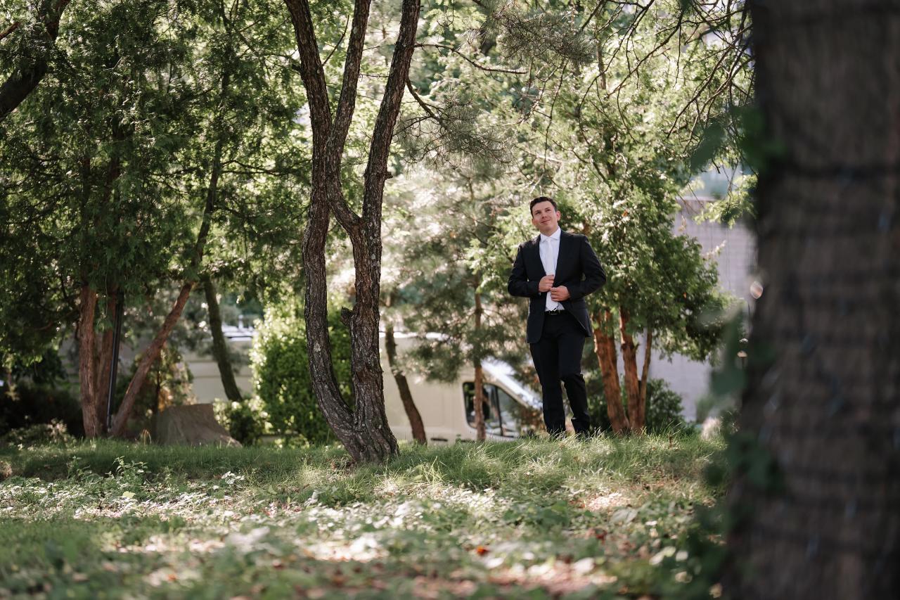 A groom in a black suit stands on grass in a garden with trees and leafy branches framing the scene, shot in soft daylight. Wedding photography at Riverview Ballroom, Cliffside Park, NJ.