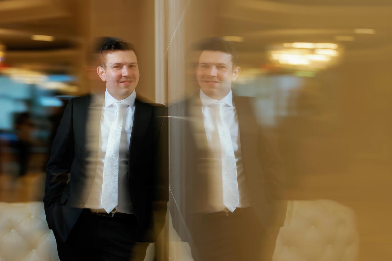 Smiling groom with a warm, reflected double-image effect near a window in soft indoor light. Wedding photography at Riverview Ballroom, Cliffside Park, NJ.