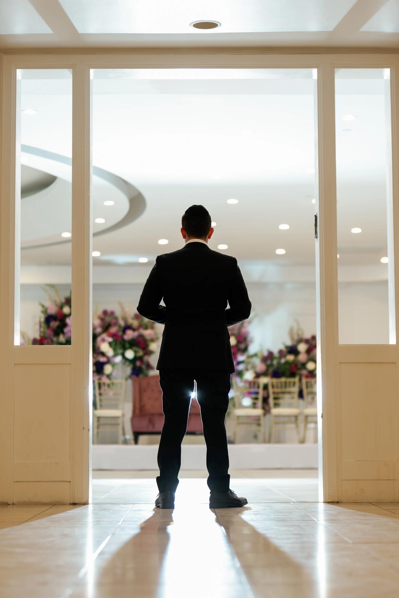 Backlit silhouette of a groom in a dark suit walking toward a bright reception space through an open doorway, with floral arrangements and chairs visible inside. Wedding photography at Riverview Ballroom, Cliffside Park, NJ.