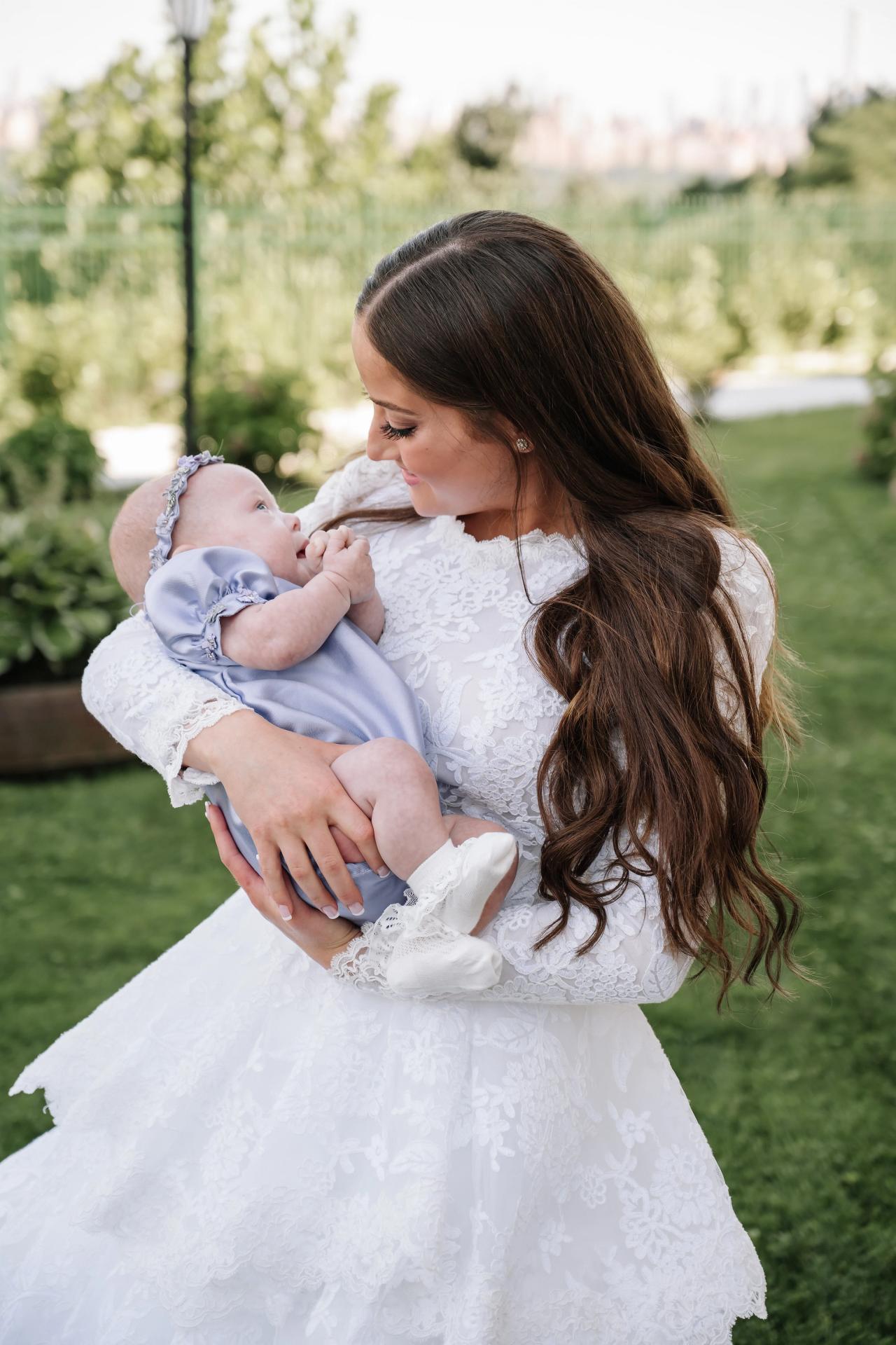 Bride in a white lace dress holding a baby outdoors, leaning in with a tender smile in a garden with soft greenery behind. Wedding photography at Riverview Ballroom, Cliffside Park, NJ.