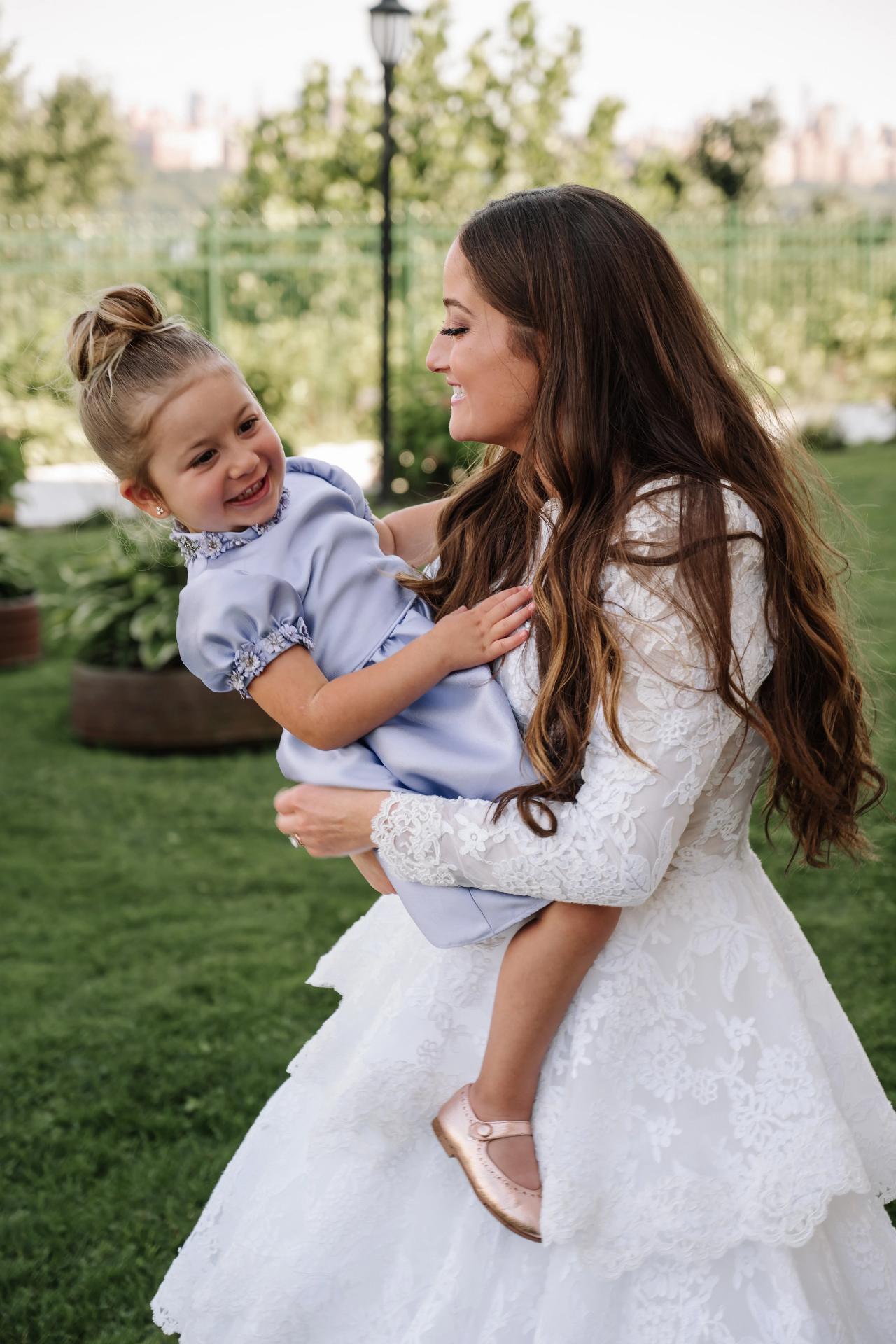 Bride holding a little girl outdoors in a garden, smiling as the child laughs and looks at her. Wedding photography at Riverview Ballroom, Cliffside Park, NJ.