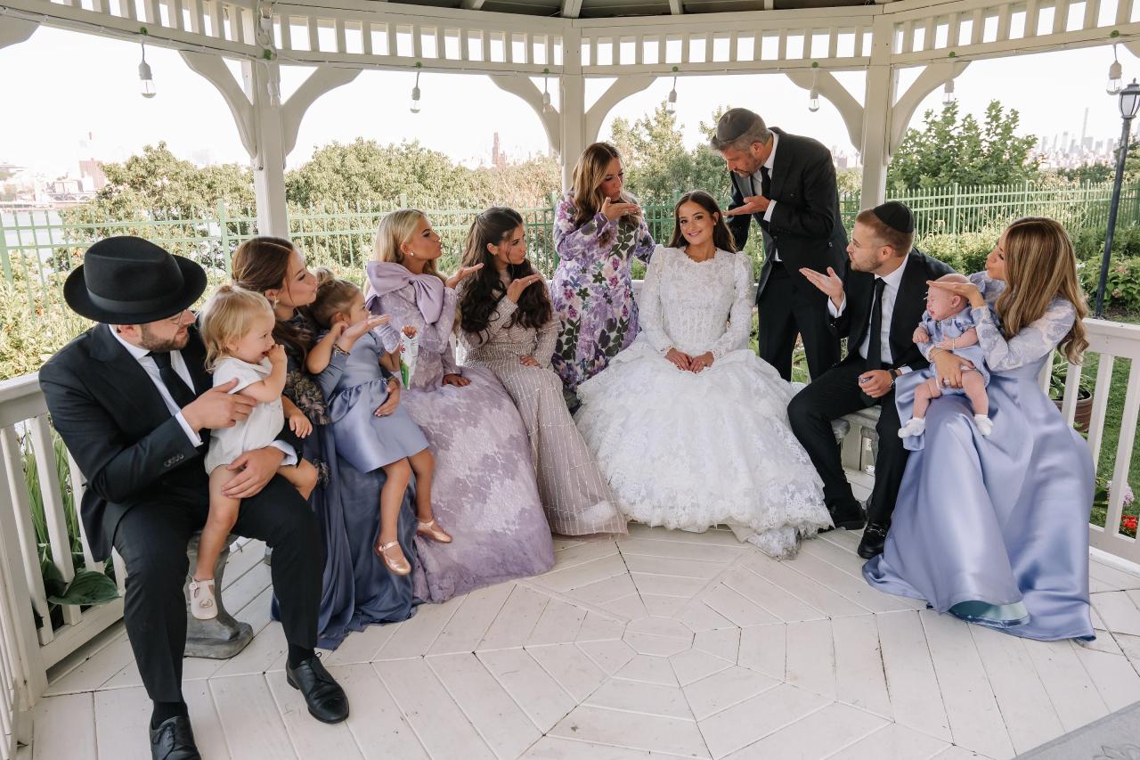Bride seated on a white gazebo patio with relatives and children gathered closely around her, guests leaning in and smiling in daylight. Wedding photography at Riverview Ballroom, Cliffside Park, NJ.